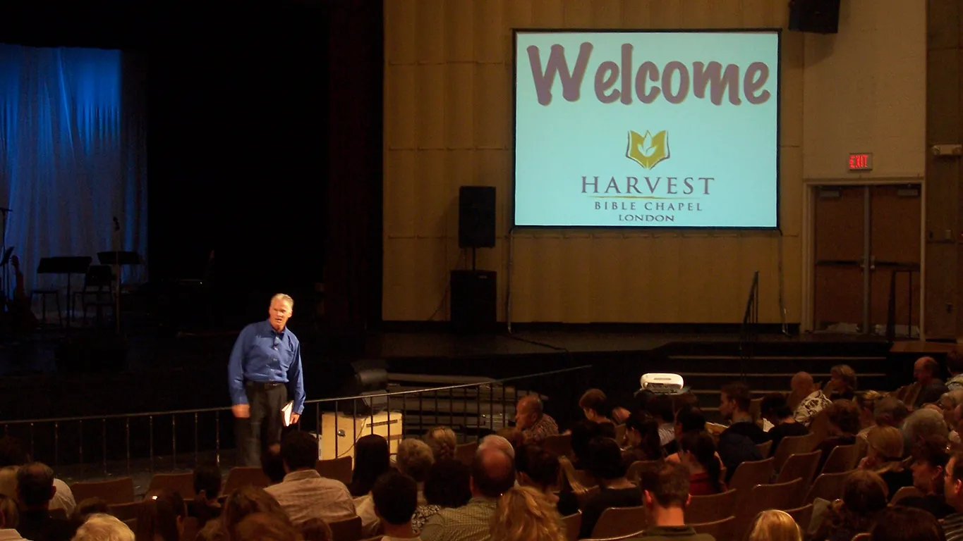 speaking on stage with screen behind with Harvest Bible Chapel logo in 2006